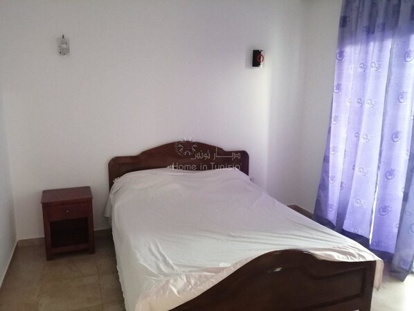 A bedroom with a bed, a nightstand, a wall-mounted light, and a curtain.