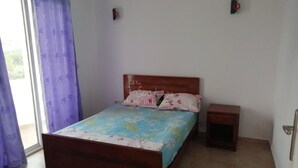 A bedroom with a wooden bed, a small bedside table, a window with purple curtains, and a view of greenery outside. - APARTMENT IN HERGLA SEA VIEW (Hergla)