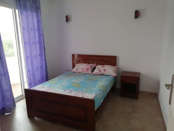 Un dormitorio con cama de madera, una mesita de noche pequeña, una ventana con cortinas moradas y vistas a la vegetación.