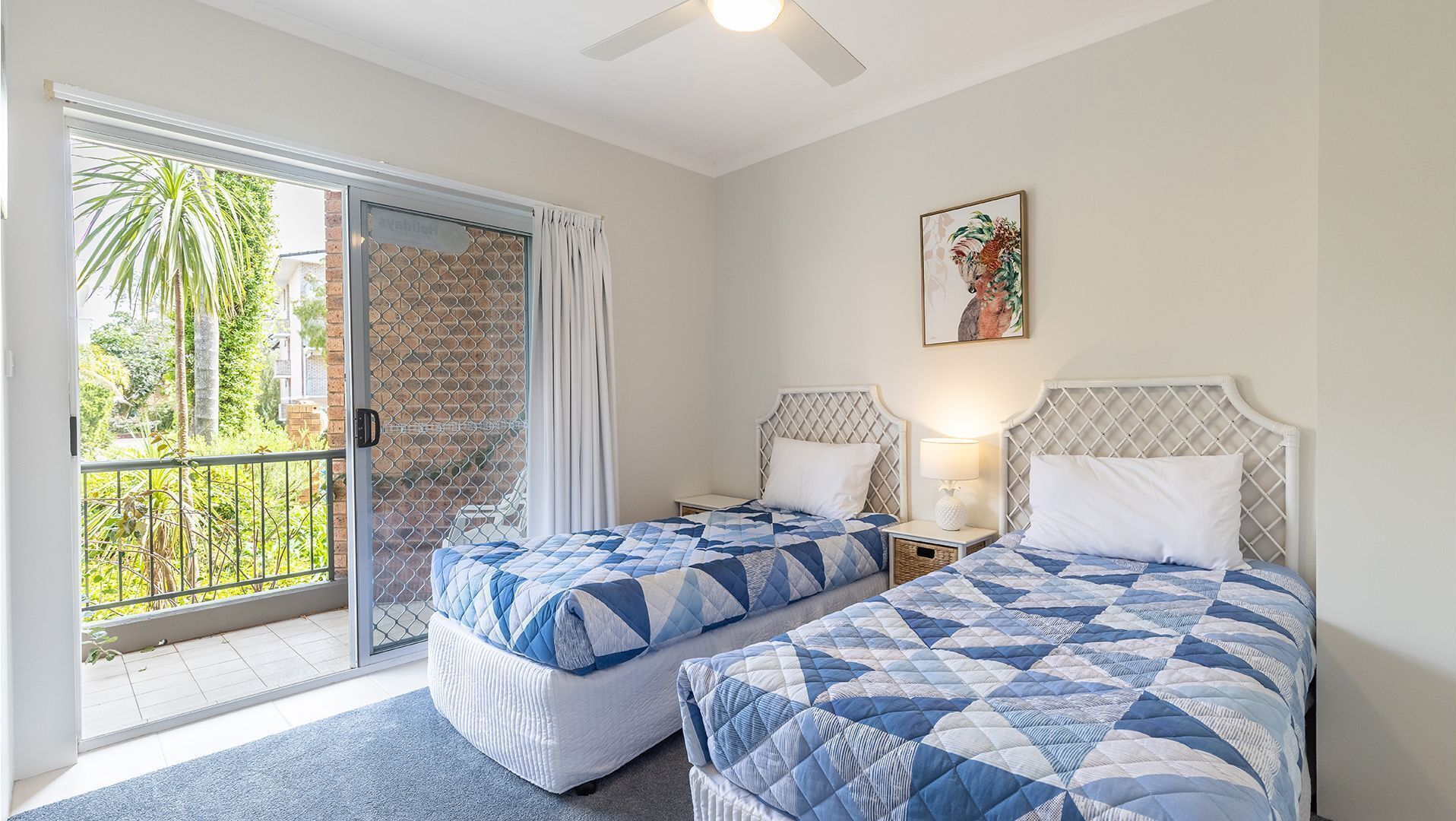 A hotel room with two beds, a balcony with a view of greenery, a ceiling fan, and a painting on the wall.
