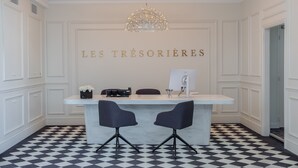 A reception area with a checkered floor, a white reception desk, two black chairs, a chandelier, and a wall with "LES TRÉSORIÈRES" written on it. - Les Trésorières (Tours)