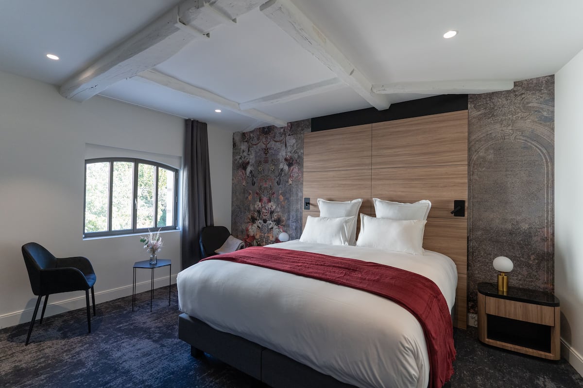 A modern hotel room with a large bed, a small table with a vase, a chair, and a window with a view of trees.