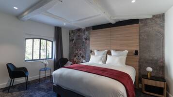 Una habitación de hotel moderna con una cama grande, una mesita con un jarrón, una silla y una ventana con vistas a árboles.