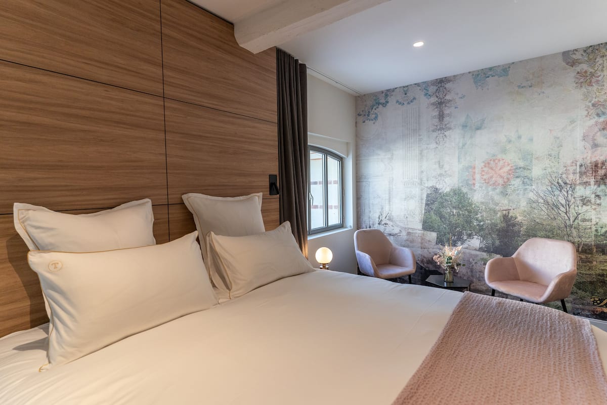 A modern hotel room with a large bed, two armchairs, a small table with a vase, and a large window.