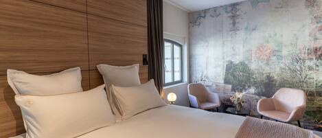 Una habitación de hotel moderna con una cama grande, dos sillones, una mesita con un jarrón y un ventanal amplio.