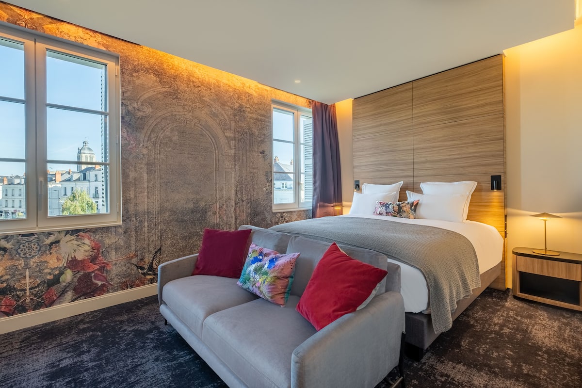 A modern hotel room with a large bed, a sofa, and a view of the cityscape through large windows.