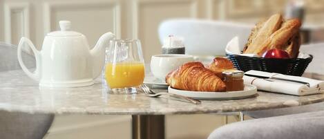 Una mesa de desayuno con cruasanes, zumo de naranja, un hervidor de agua y una cesta de pan.