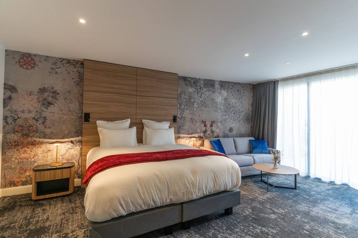 A modern hotel room with a large bed, a sofa, a small table, and a decorative wall.