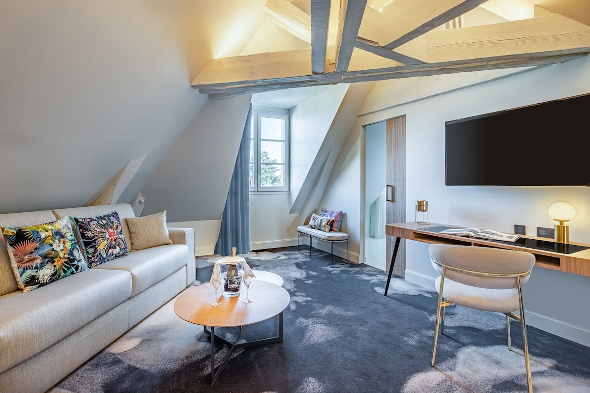 A modern loft bedroom with a sloped ceiling, a large window, a flat-screen TV, a sofa, a coffee table, and a desk with a lamp.