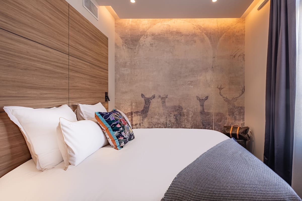 A modern hotel room with a large bed, white and colorful pillows, a headboard with wood paneling, a textured wall with deer silhouettes, and a window with curtains.