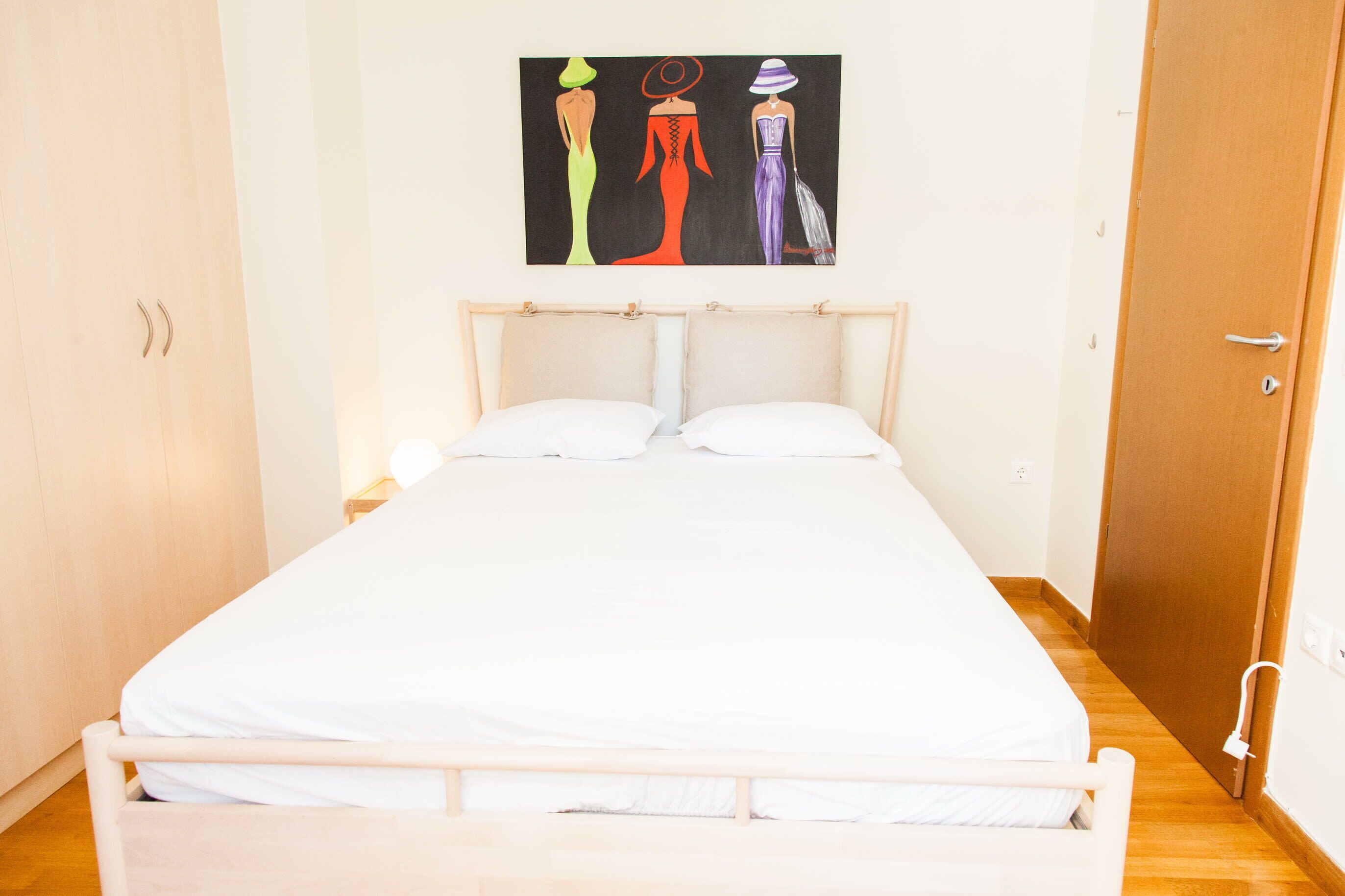 A single bed with a wooden headboard, white bedding, and a painting of three figures on the wall.