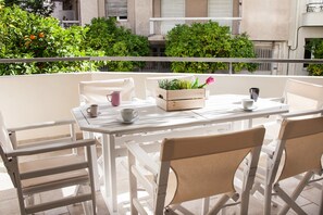 A balcony with a white table and chairs, a wooden planter box with plants and a pink flower, and a view of green trees and buildings. - Sifris Apartment in the Heart of Athens (Athens)