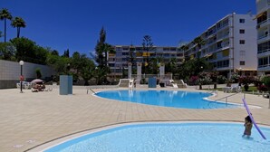 Une piscine où un enfant joue, entourée d’un immeuble à plusieurs étages doté de balcons, sous un ciel bleu clair.