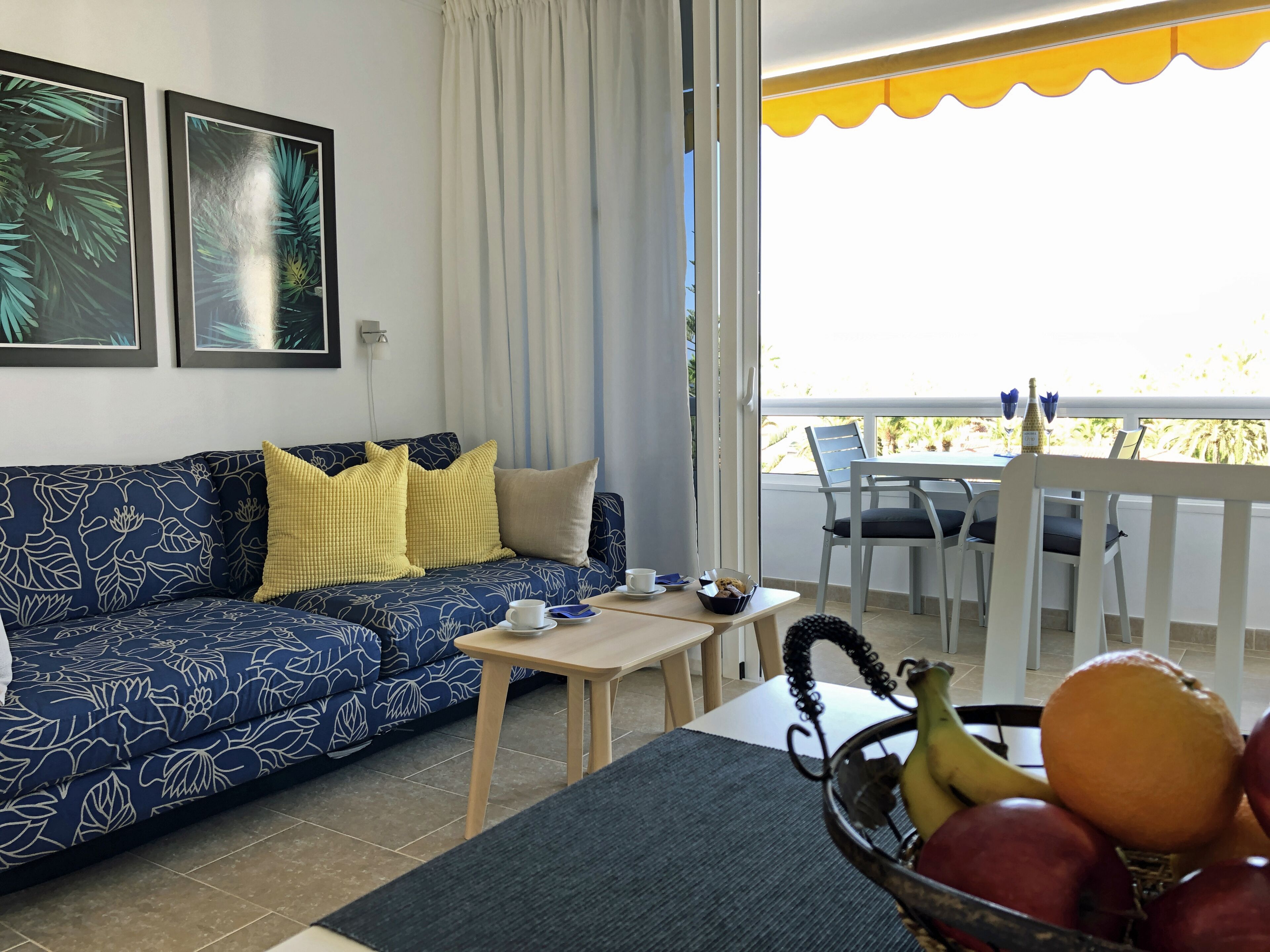 Ein Wohnzimmer mit blauem Sofa, gelben Kissen, einem kleinen Tisch mit Tasse und Untertasse sowie einem Obstkorb.