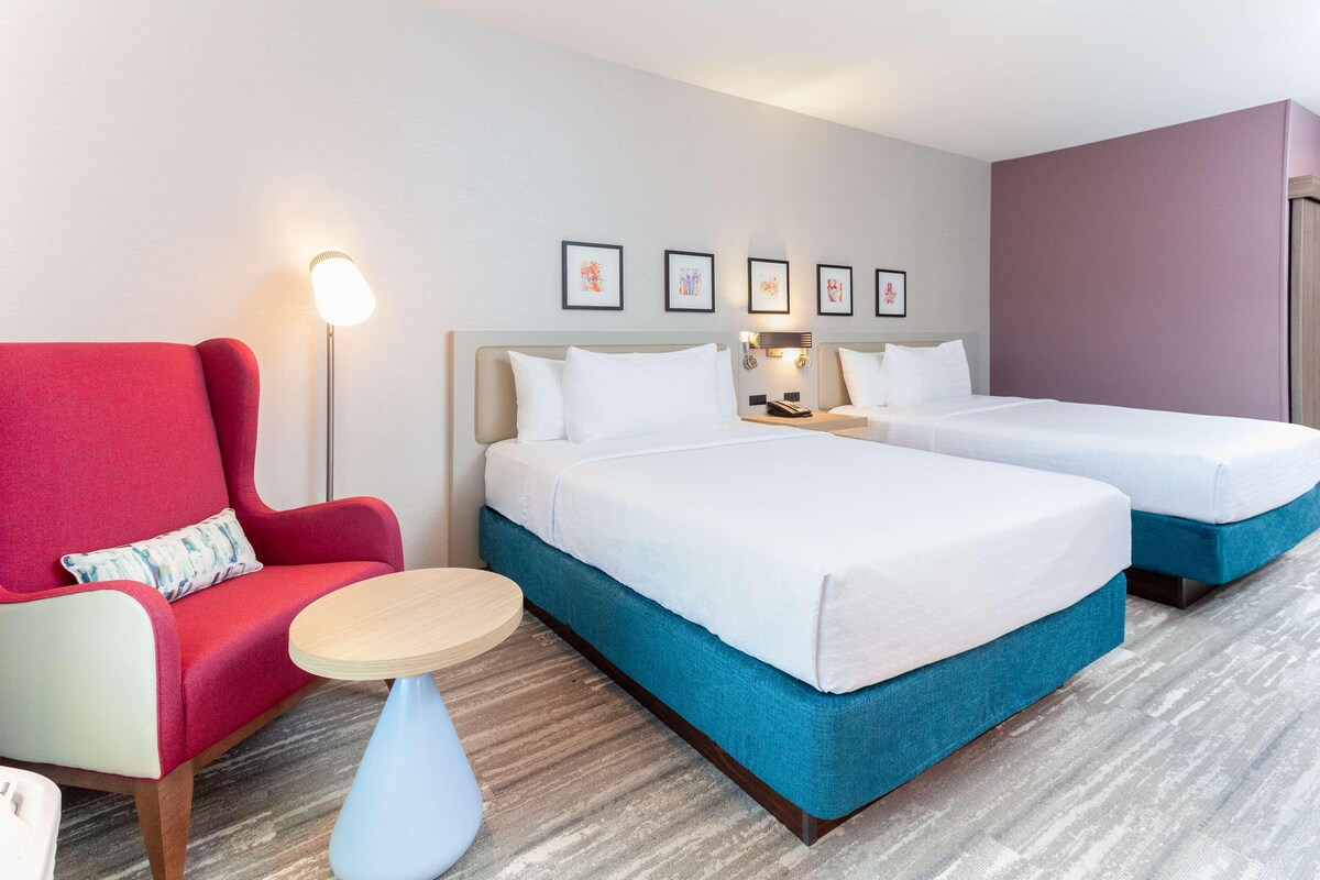 A hotel room with two beds, a red armchair, a small round table, and a floor lamp.