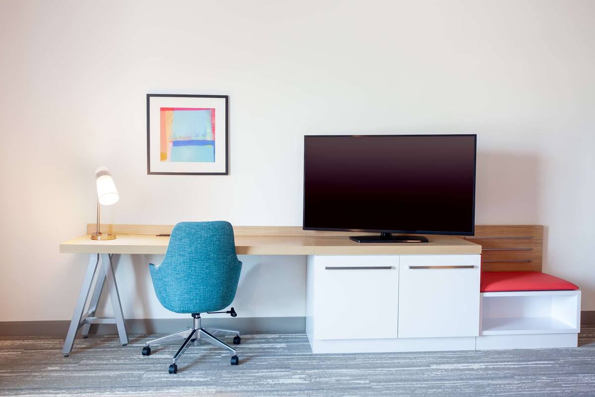 A modern hotel room with a desk, a blue office chair, a television, and a framed picture on the wall.