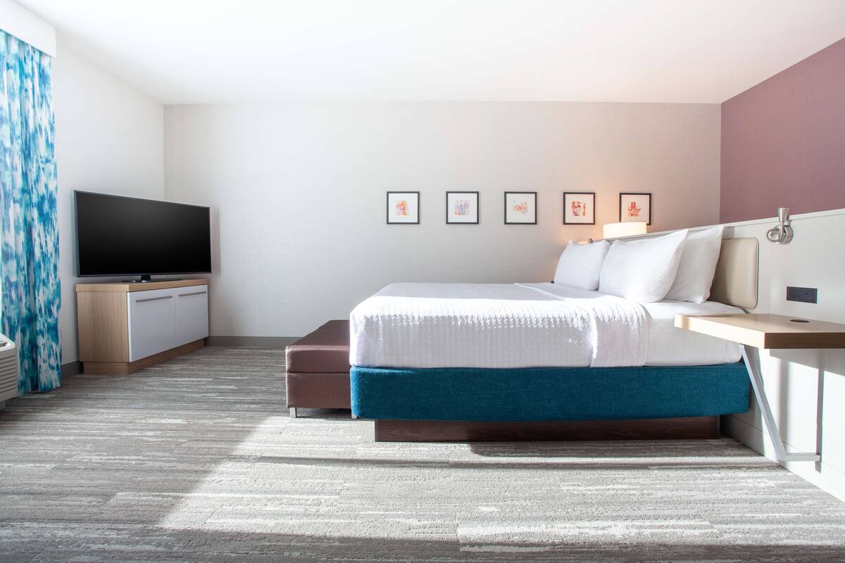 A modern hotel room with a bed, bedside table, TV, and artwork on the wall.