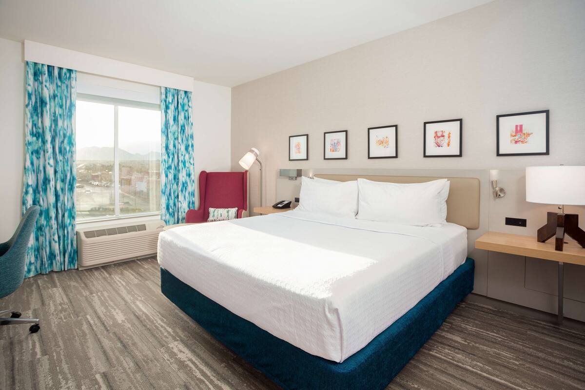 A hotel room with a large bed, a desk with a lamp, a chair, and a window with curtains.