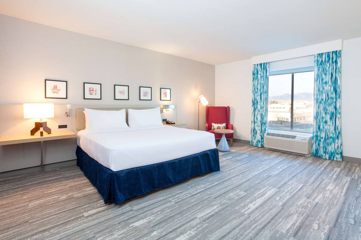 A hotel room with a large bed, a desk, a chair, a window with curtains, and framed pictures on the wall.