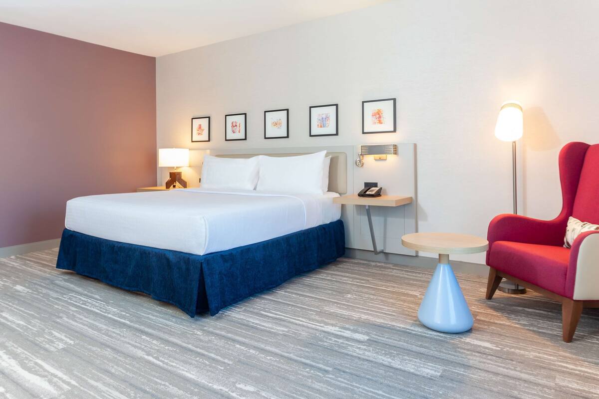 A hotel room with a bed, a nightstand, a lamp, a chair, and framed pictures on the wall.