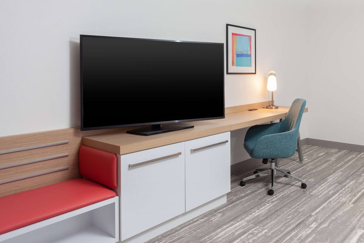 A modern room with a flat-screen TV on a wooden desk, a red cushioned bench, a teal office chair, and a framed picture on the wall.
