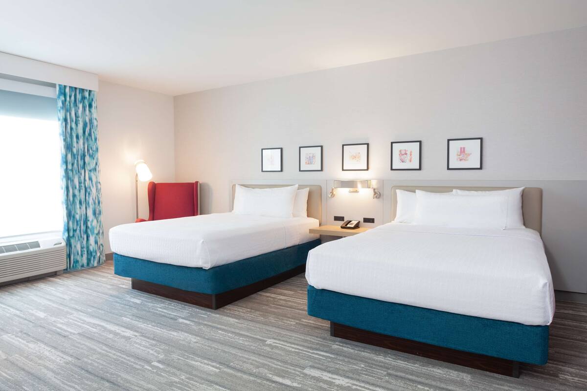 A hotel room with two beds, a window with curtains, a bedside table with a lamp, and framed pictures on the wall.
