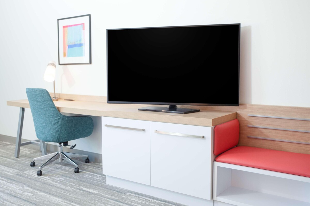 A modern office desk with a flat-screen TV, a teal office chair, a red cushioned bench, and a framed picture on the wall.