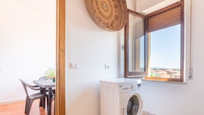 A small room with a washing machine, a table with chairs, a potted plant, and a window with a view of buildings.