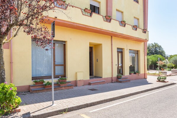 A yellow building with multiple windows and doors, some with potted plants. There are steps leading to the entrance and a sidewalk in front. - Vacation Home da Placida "S'Archittu" (Suni)