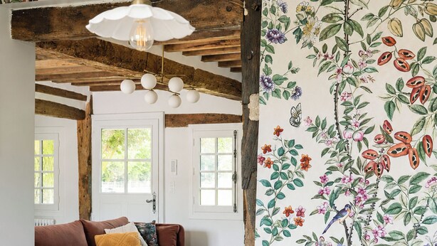 Une pièce avec un plafond en bois, un canapé, une table basse, un plateau sur lequel se trouve un vase de fleurs, et un mur recouvert d’un papier peint à motifs floraux.