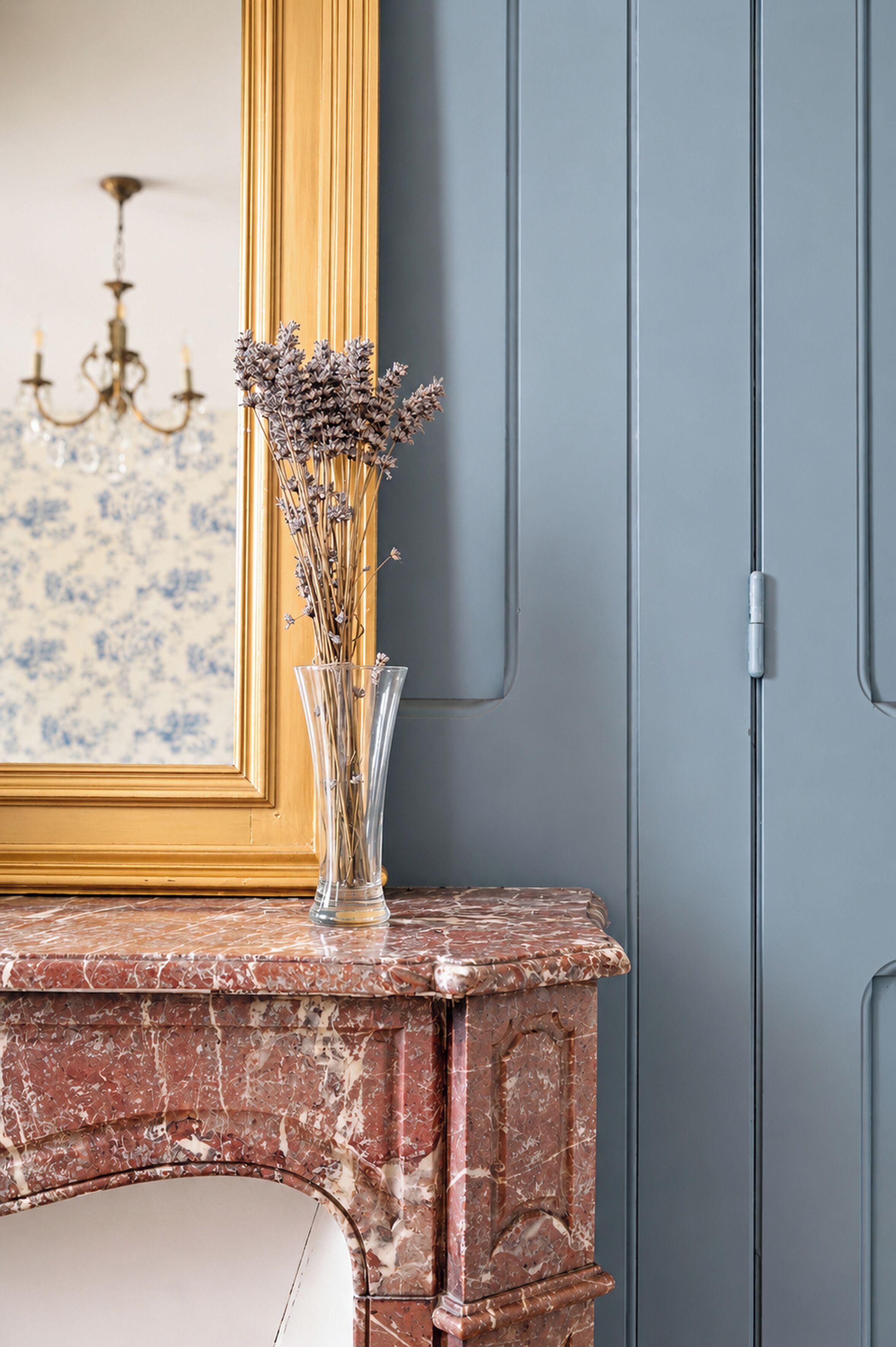 A marble fireplace with a vase of dried flowers, a framed mirror, and a chandelier in the background.