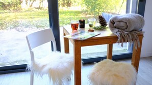 A wooden table with a magazine, a candle, and a pine cone. There are two white furry chairs and a window with a view of greenery. - A cozy n'est by the lake, the Azur studio (Sévrier)