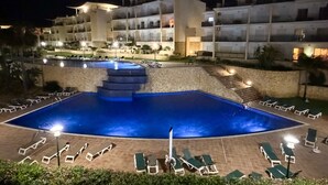 Una piscina con escalones de acceso, rodeada de sillas de descanso y un edificio con varios balcones.