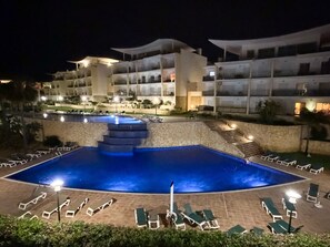 A swimming pool with steps leading into it, surrounded by lounge chairs and a building with multiple balconies.