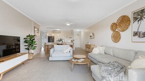 Ein modernes Wohnzimmer mit Sofa, Couchtisch und Fernseher. Im Hintergrund ist eine Essecke zu sehen, mit einer angrenzenden Küche.