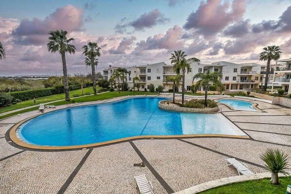 A large, curved swimming pool with a pebble-lined deck, surrounded by palm trees and a building with multiple balconies.