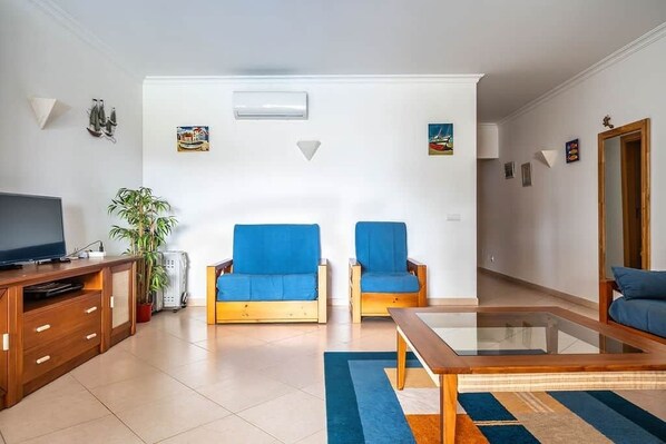 Una sala de estar moderna con dos sillones azules, una mesa de centro de madera, un televisor sobre un mueble de madera y una planta en maceta.