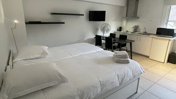 Una cama bien hecha con sábanas blancas, una mesita de noche con lámpara, un televisor de pantalla plana en la pared, una pequeña mesa de comedor con sillas y una cocina con fregadero y estufa.