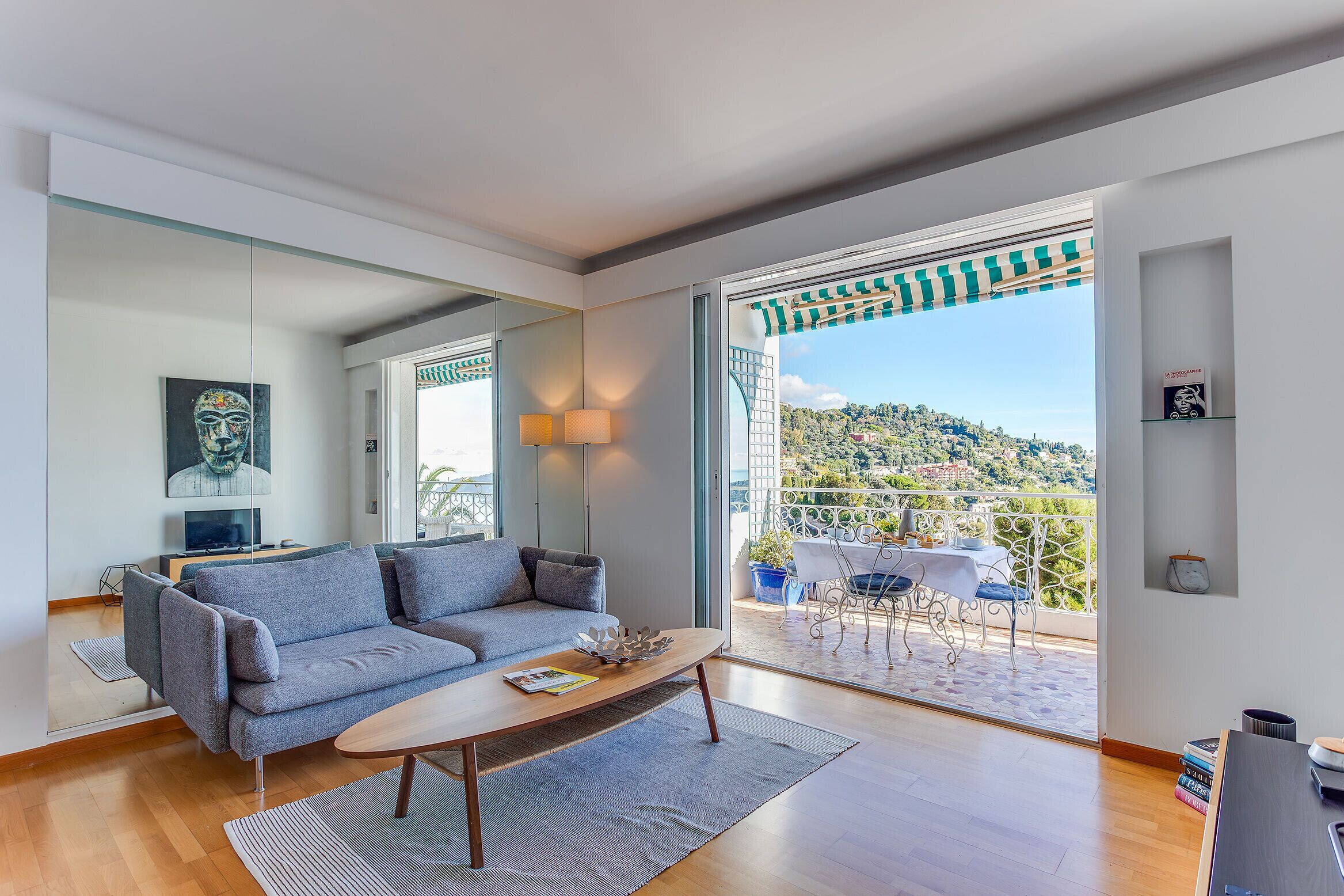 A modern living room with a grey sofa, a wooden coffee table, and a balcony with a view of a hillside.