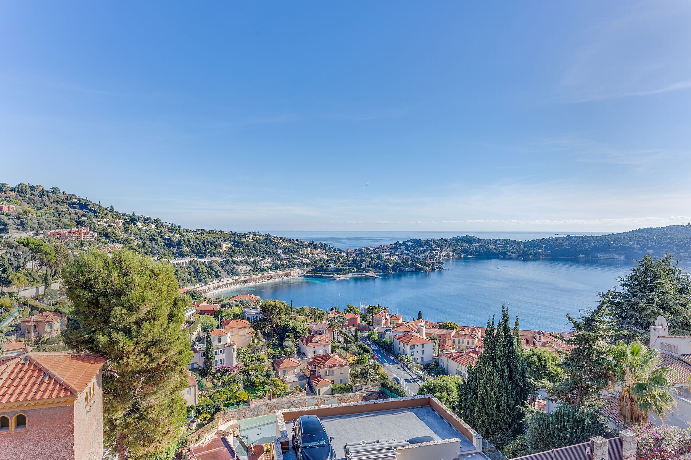 A coastal town with terraced houses, a clear blue sea, and a prominent tree in the foreground.