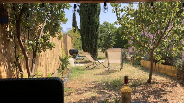 Un espace patio avec une table en bois, deux verres à vin et une bouteille de vin. On aperçoit des chaises et un barbecue à l’arrière-plan.