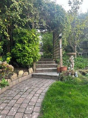 Una escalera de piedra que sube hacia un portón de madera, rodeada de abundante vegetación y plantas en macetas.