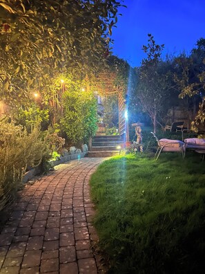 Un sendero pavimentado que conduce a un espacio cubierto con escalones, rodeado de vegetación y mobiliario exterior.