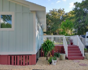 A light blue building with a red base, white railings, and a red staircase leading to a porch area with potted plants.