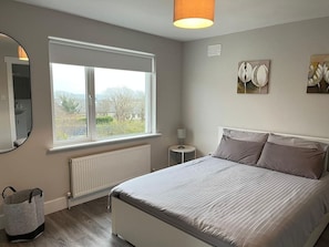 A bedroom with a large window, a bed with grey bedding, a bedside table, a lamp, and a radiator. - Bay Area House, Spiddal (Spiddal)