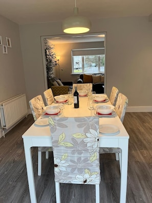 A dining area with a table set for four, a bottle of wine, and a view of the outside through a window. - Bay Area House, Spiddal (Spiddal)