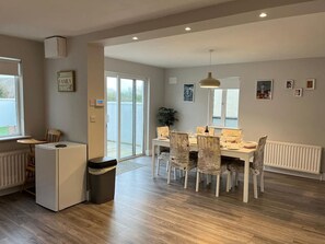 A dining area with a table and chairs, a refrigerator, a trash bin, and a radiator. - Bay Area House, Spiddal (Spiddal)