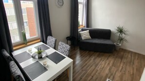 Uma sala de estar com uma mesa branca, cadeiras estampadas, um sofá escuro e um vaso de planta.