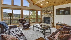 Ein geräumiges Wohnzimmer mit Ledersofas, einem Kamin, einer Wand aus Naturstein und großen Fenstern mit Blick ins Freie.