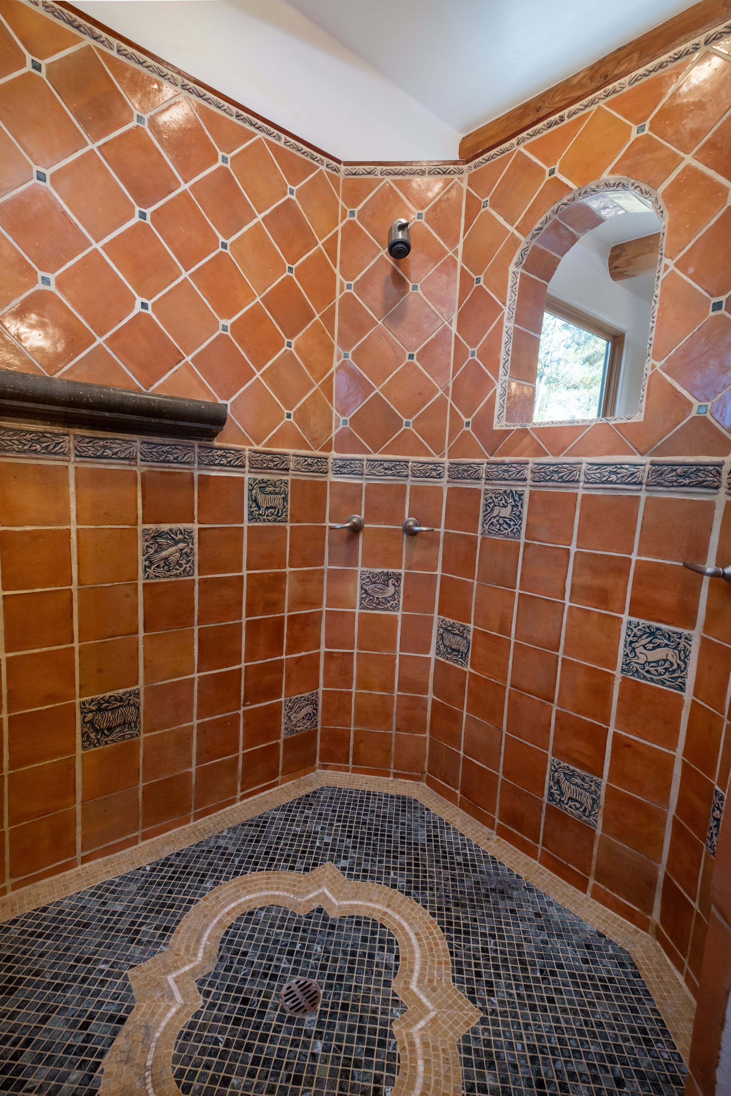 Een douche in een hoek, met terracotta tegels en decoratieve patronen, een klein raam en een metalen douchekop.